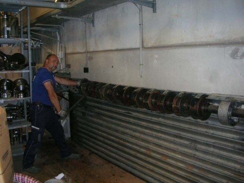 Uomo che installa un lungo rullo metallico su una parete con tubi metallici sottostanti. Gli scaffali con gli oggetti sono sulla sinistra.