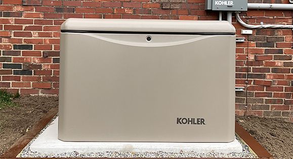 Tan KOHLER generator on a concrete pad, next to a brick wall with electrical components.