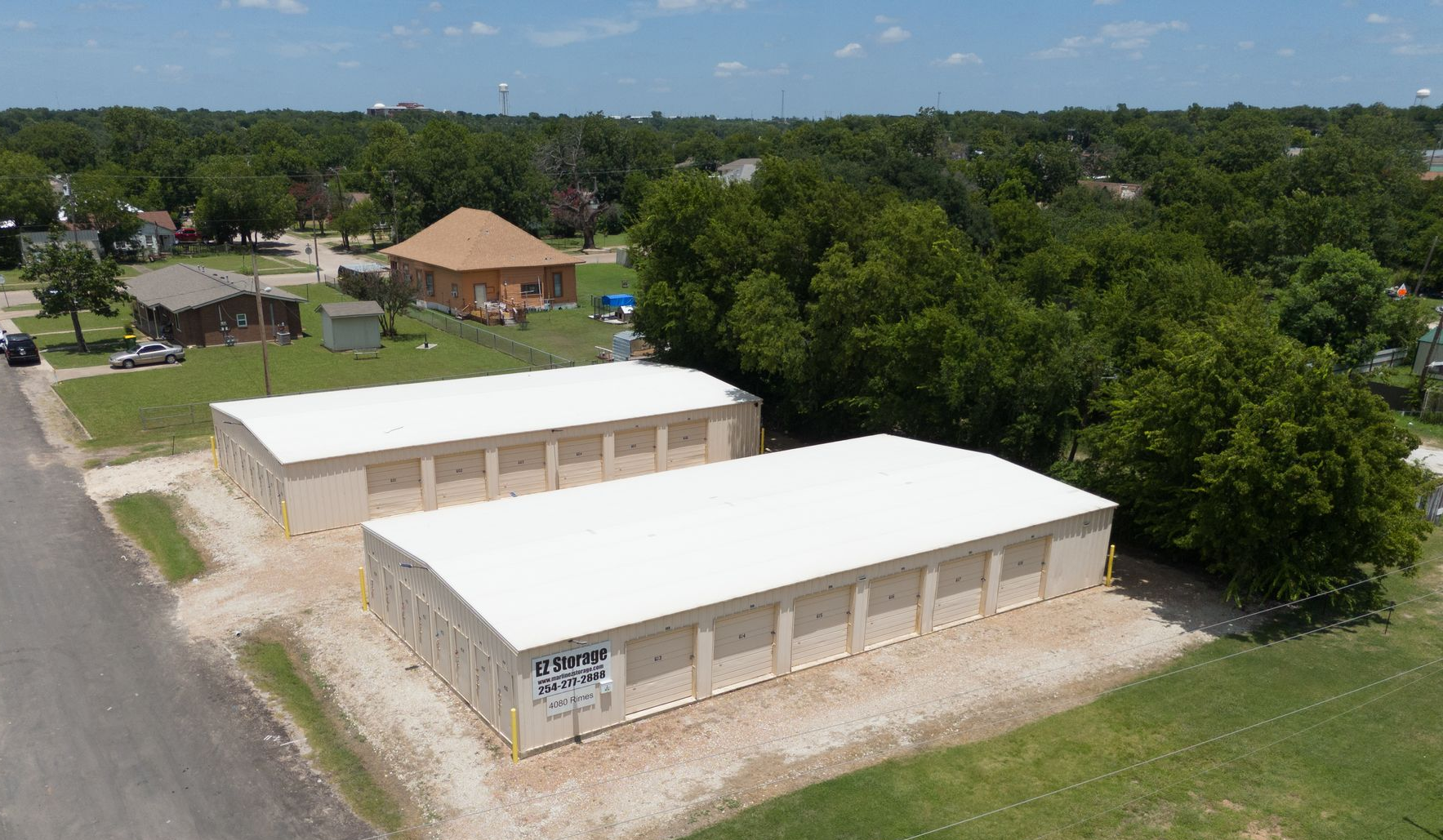 An aerial view of two beige, flat-roofed storage unit buildings next to a road, grass, and trees in a residential area.
