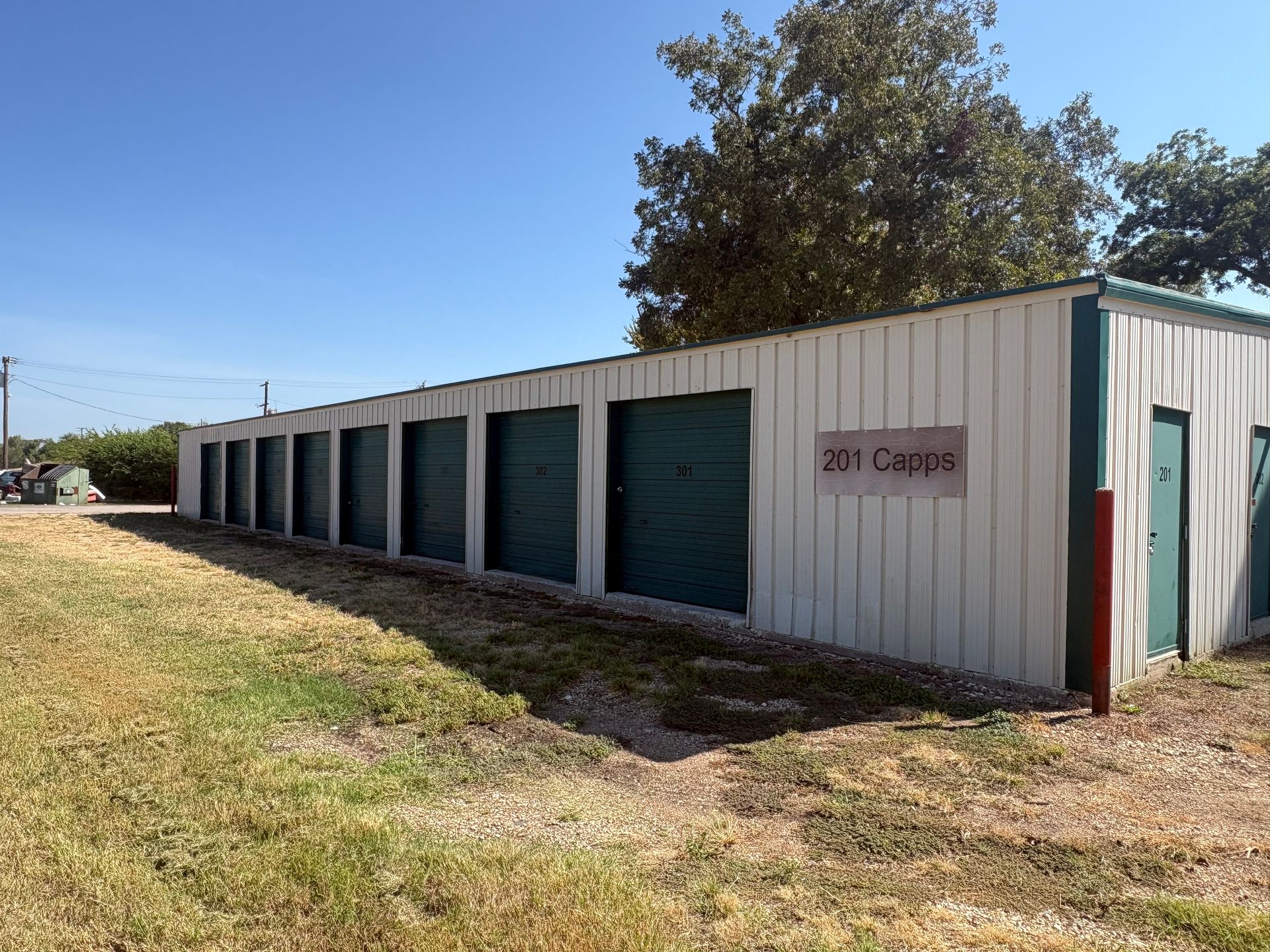 Row of green storage unit doors in a beige metal building on a grassy roadside under a blue sky