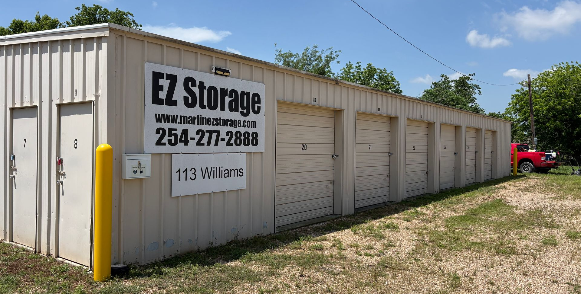 Row of beige EZ Storage units with a sign, bordered by grass and trees.
