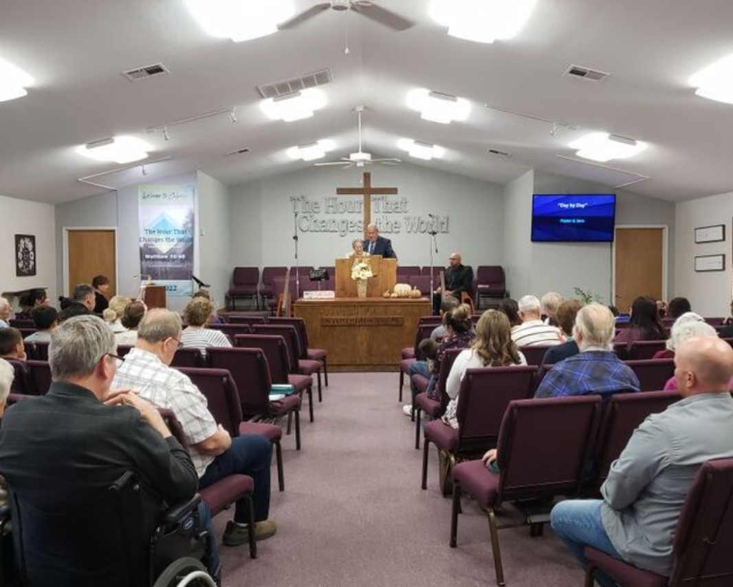 Congregation seated in a small church hall, listening to a speaker at the front beneath a cross.