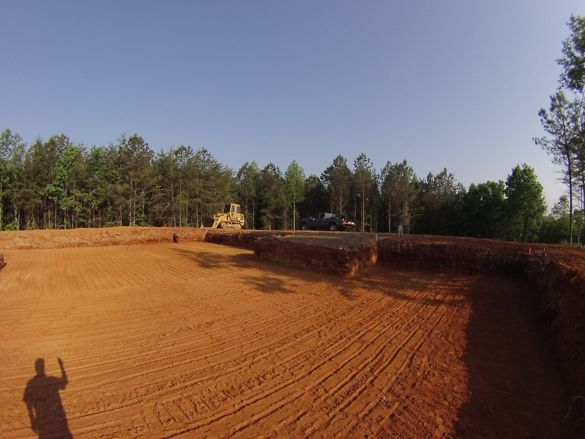 Construction site with bulldozer shaping red dirt beside trees under a clear sky
