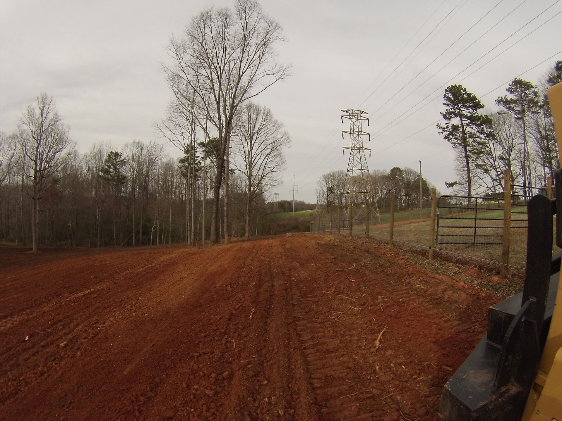 Reddish dirt road curving through a wooded rural area beside a fence and gate under an overcast sky