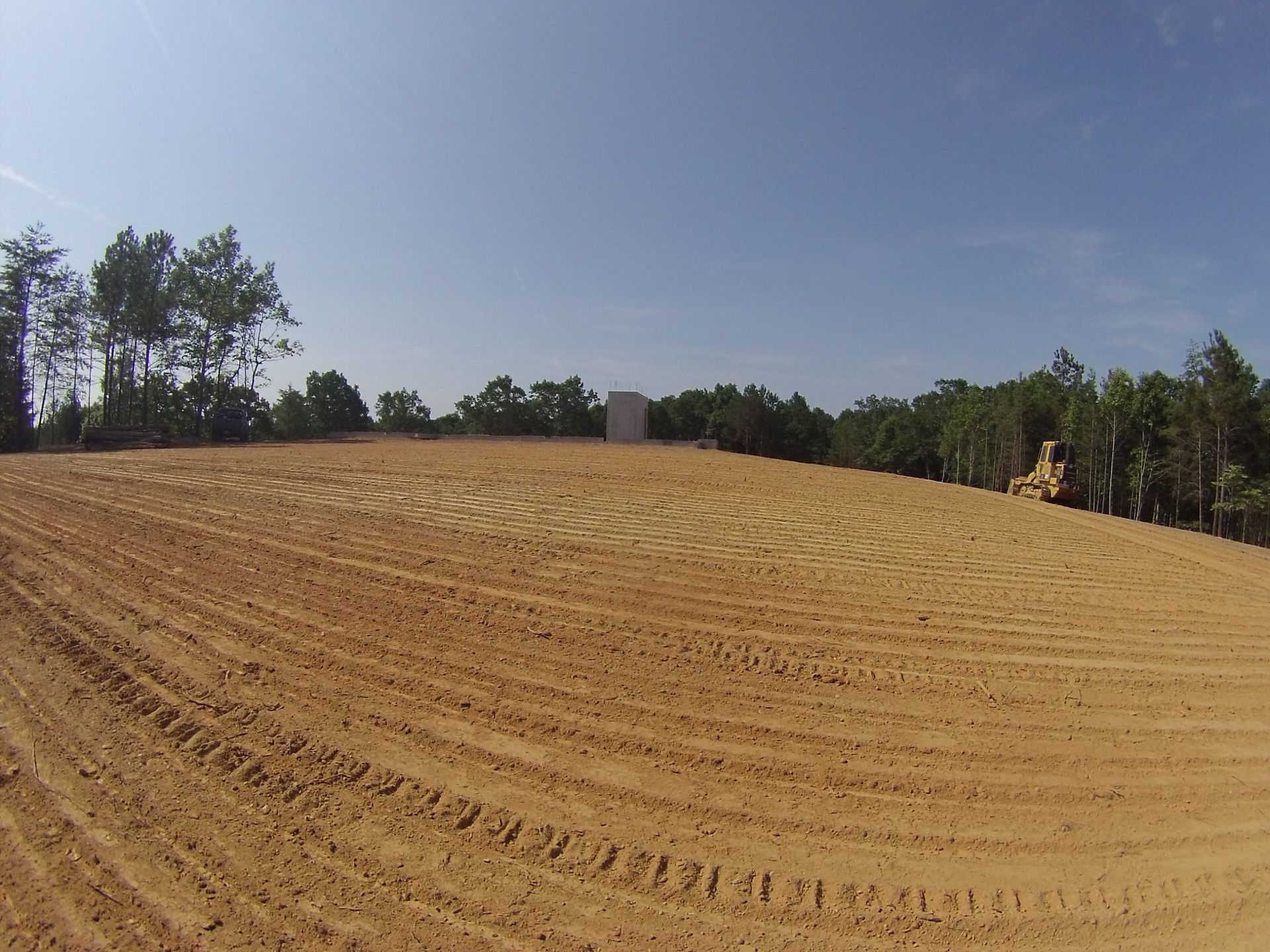 Wide dirt field under a blue sky, with trees along the edge and a person in yellow at the far right.