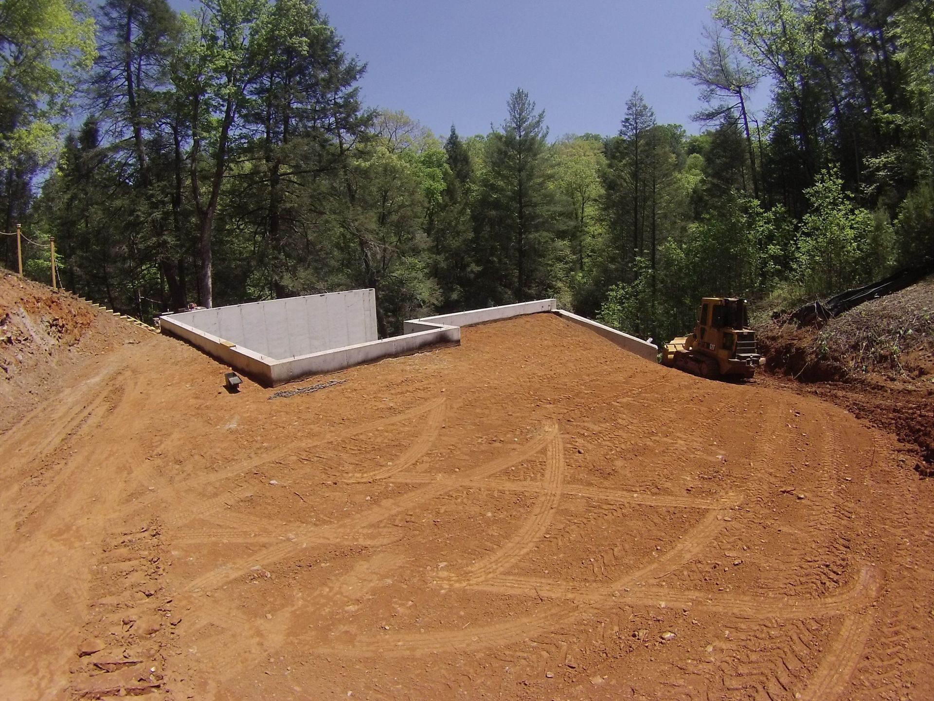Cleared dirt lot with a concrete foundation, trees behind, and a small bulldozer at right