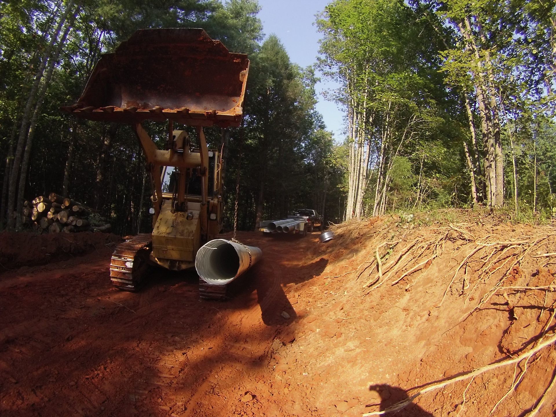 Small bulldozer on a red dirt road beside a large concrete pipe in a wooded construction site