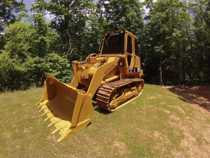 Yellow bulldozer on a grassy dirt path beside trees, viewed from the front-left.