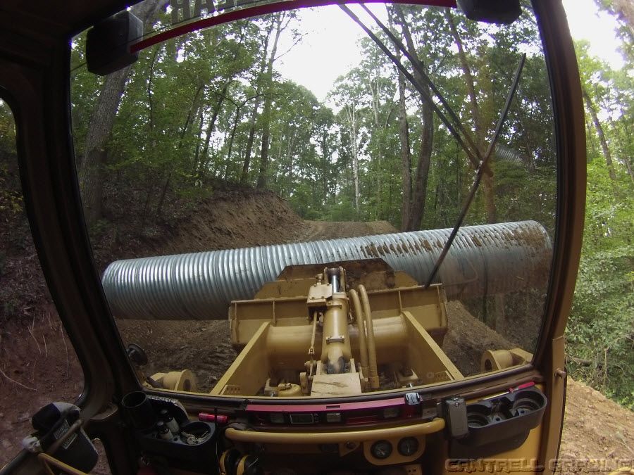 Cab view of a yellow bulldozer pushing a large corrugated pipe through a wooded construction site