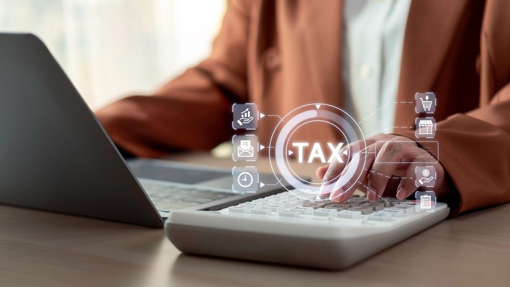 Hands using a calculator beside a laptop, with a glowing “TAX” icon overlay.