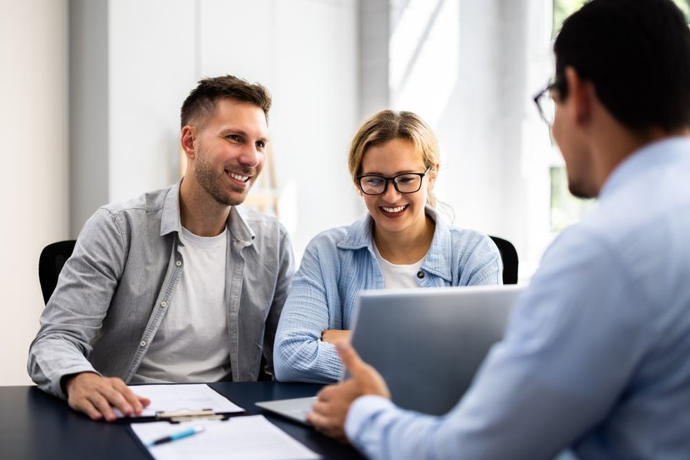 Three coworkers smiling and talking during a meeting at a laptop, with papers on the table.