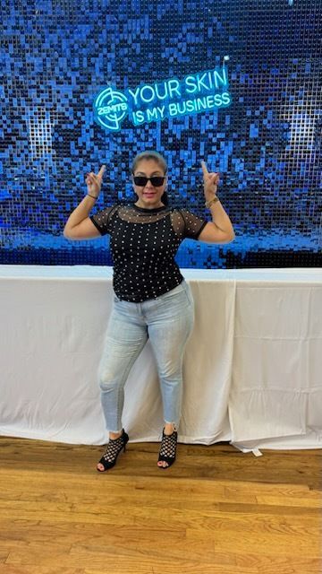Woman in black top and jeans, pointing up, in front of a blue sequined wall with a neon sign.