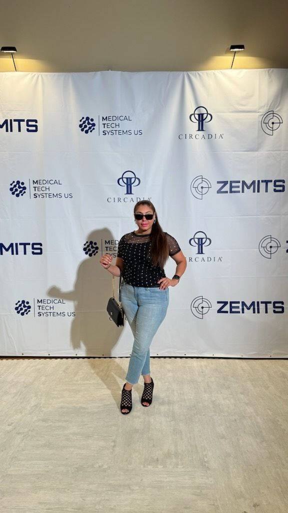 Woman poses in front of a backdrop, wearing sunglasses, black top, and jeans, holding a small purse.
