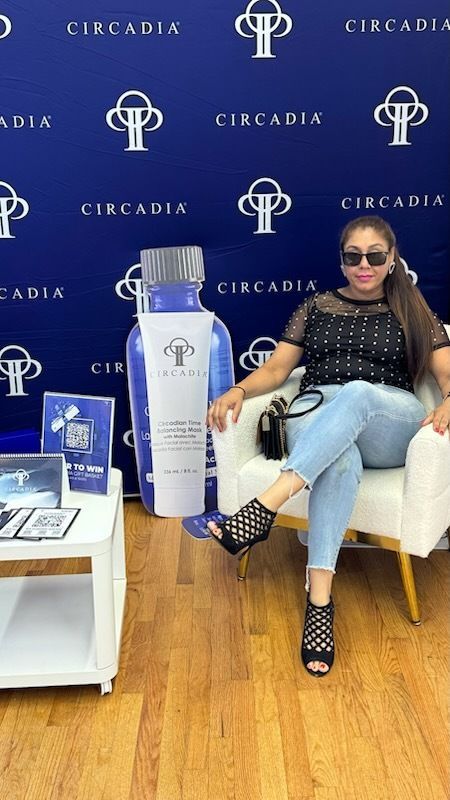 Woman in sunglasses poses with Circadia products. Blue backdrop, seated in chair, wearing jeans, black top, heels.