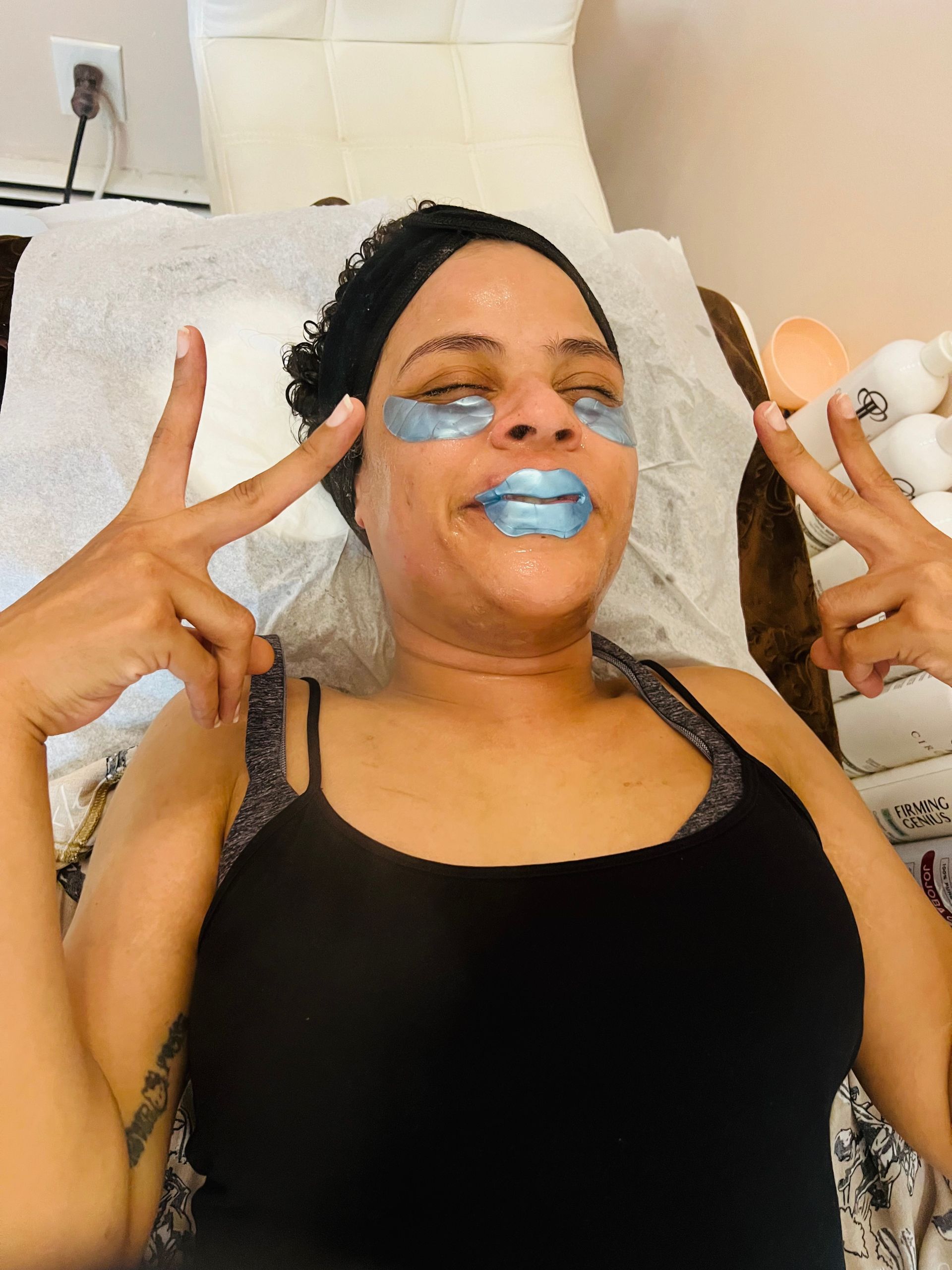 Woman with eye and lip masks making peace signs, lying on a spa bed.