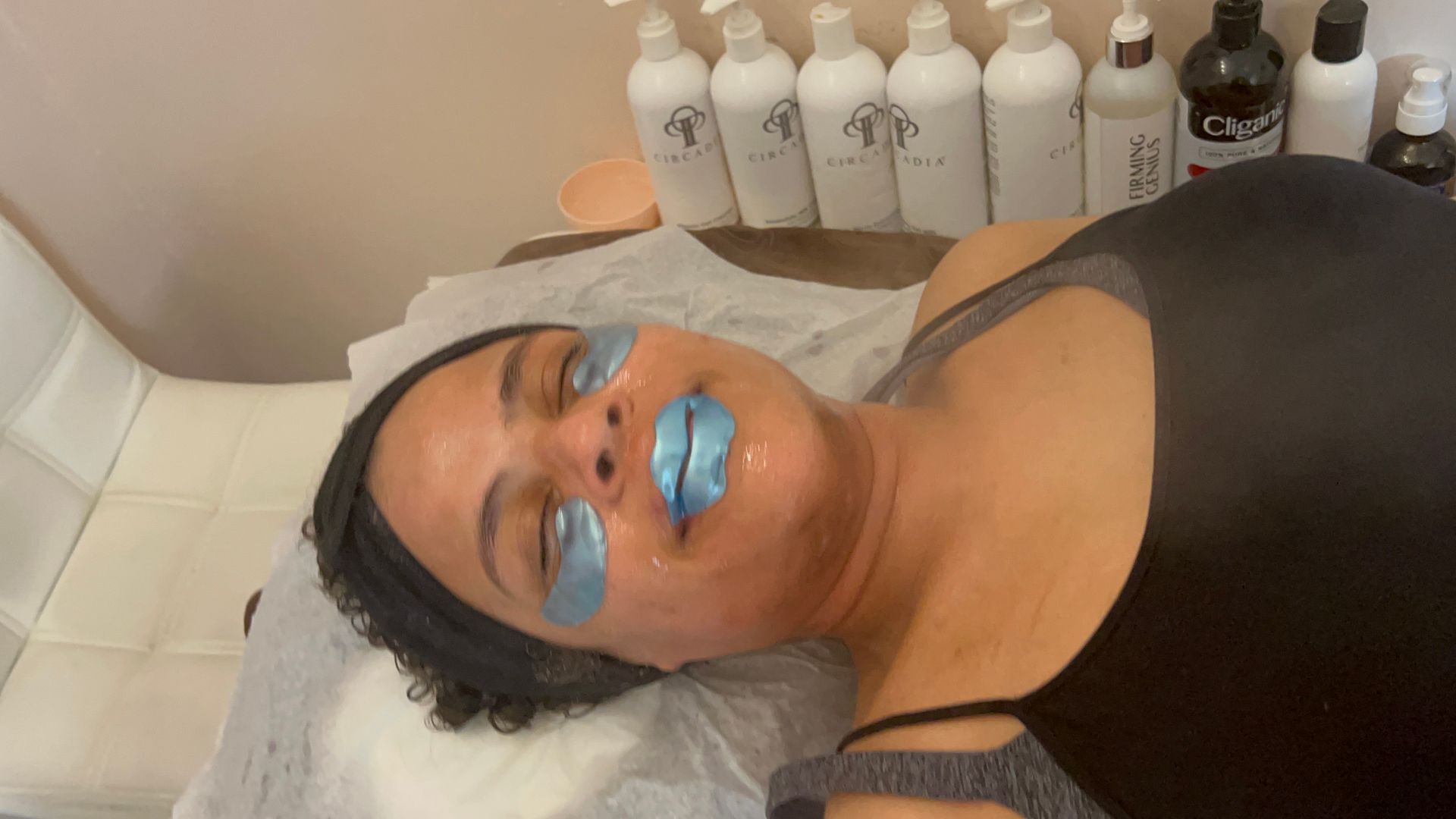 Person reclining with blue eye and lip masks on, in a treatment room. Skincare products are in the background.