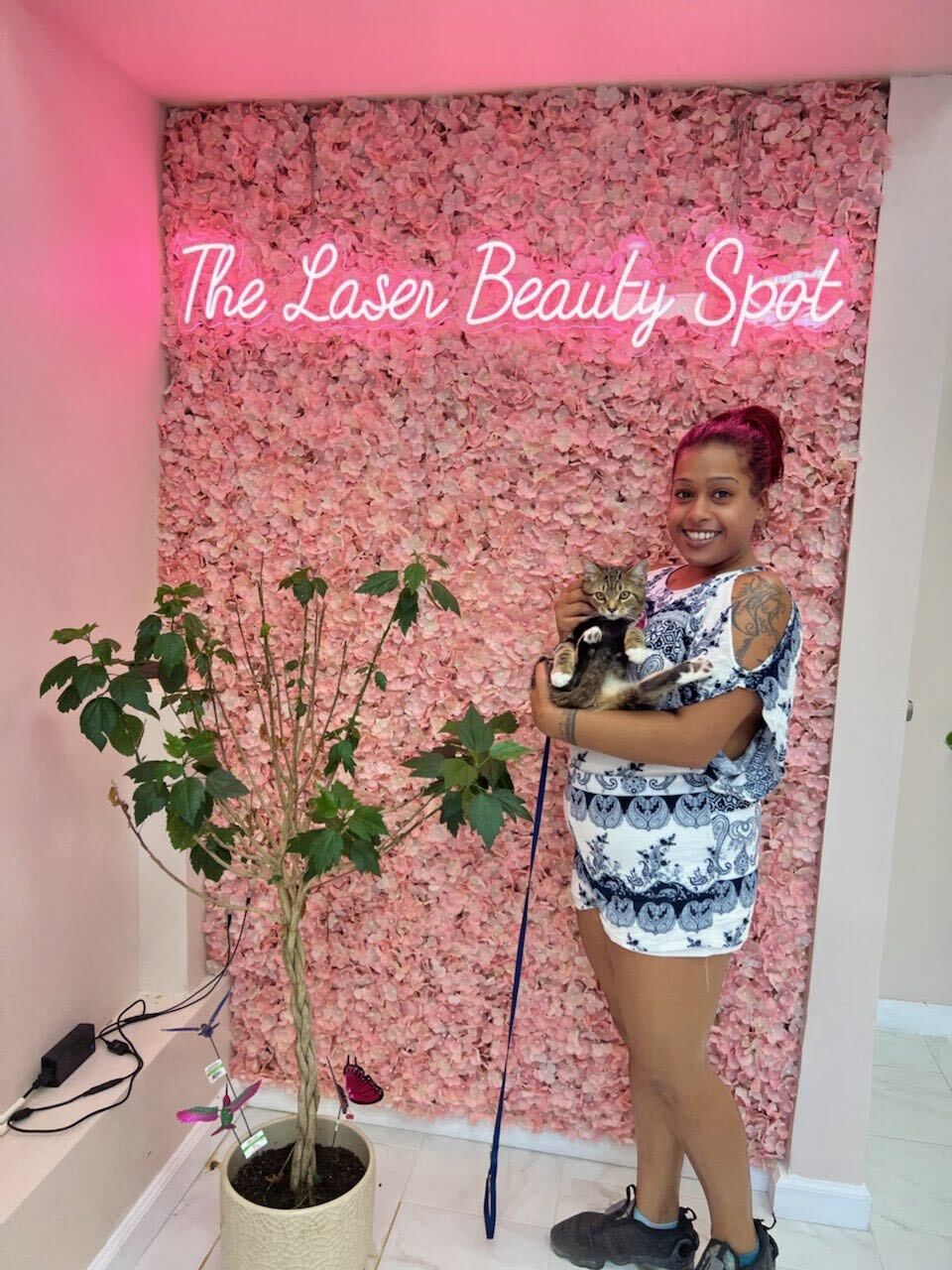 Woman holding a cat in a beauty spot, in front of a pink floral wall, and neon sign.