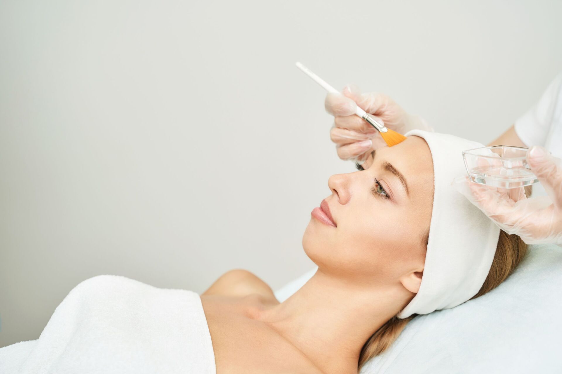 Person receiving a facial peel treatment; a cosmetic professional applies a chemical solution with a brush.