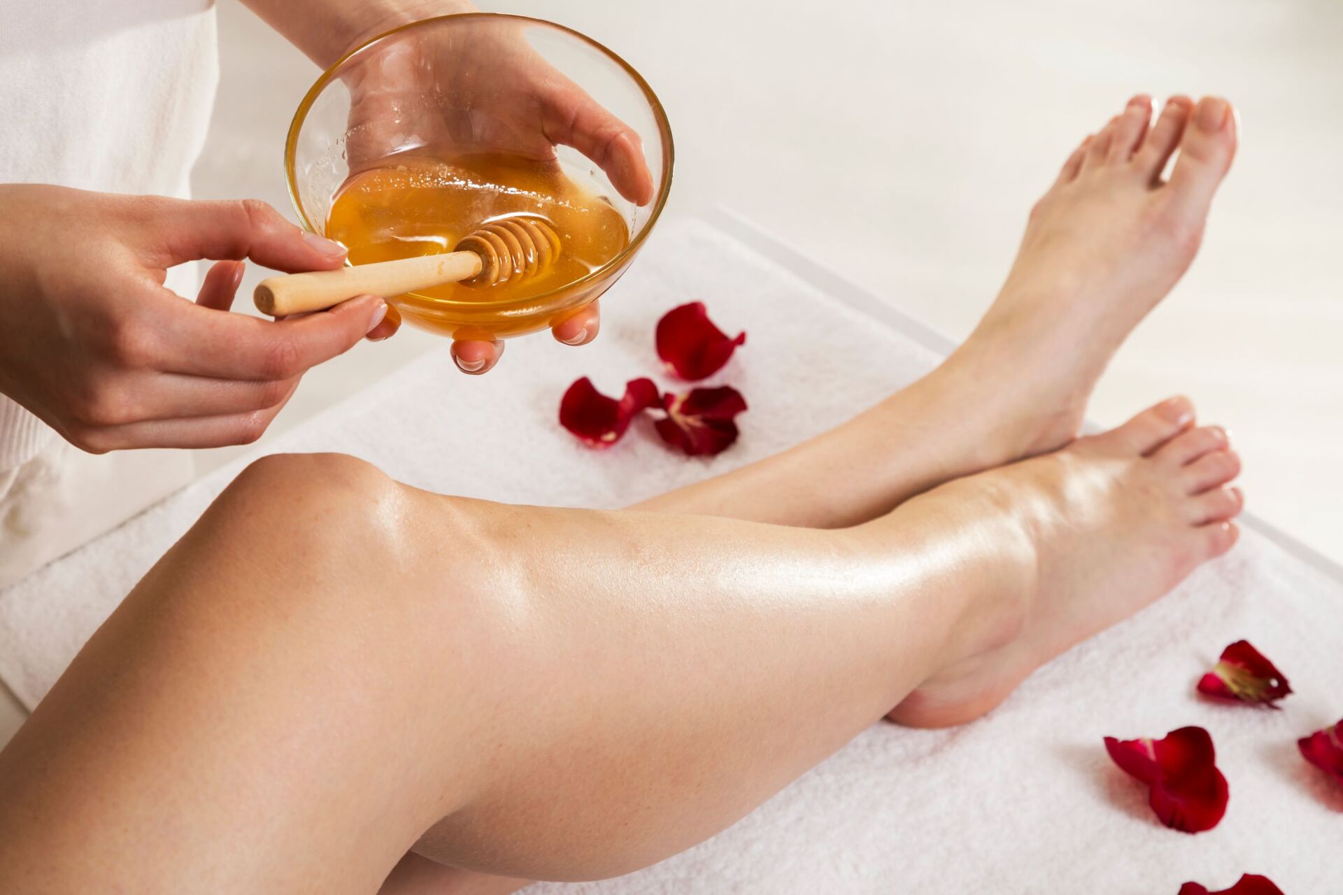 Legs being waxed, honey and applicator in hand, on a white towel with rose petals.