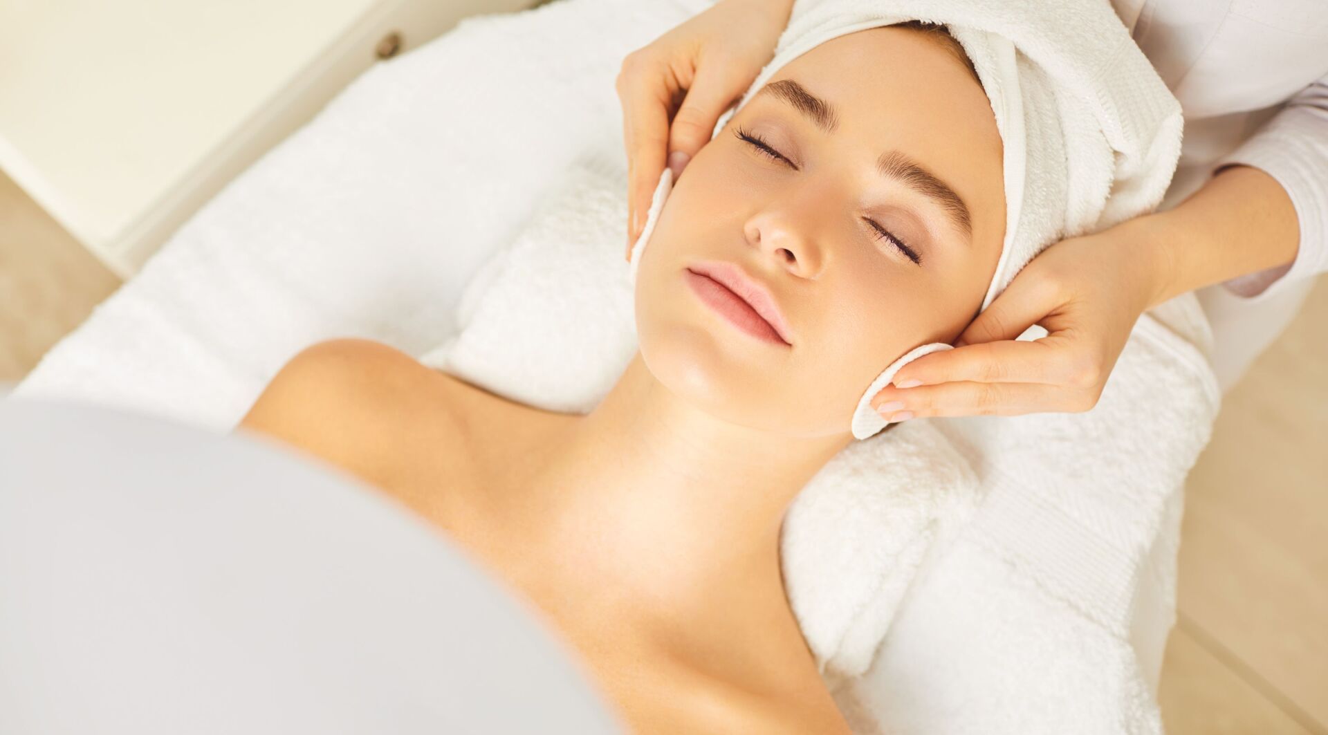 Woman receiving facial treatment in spa, eyes closed, with towels and hands on face.