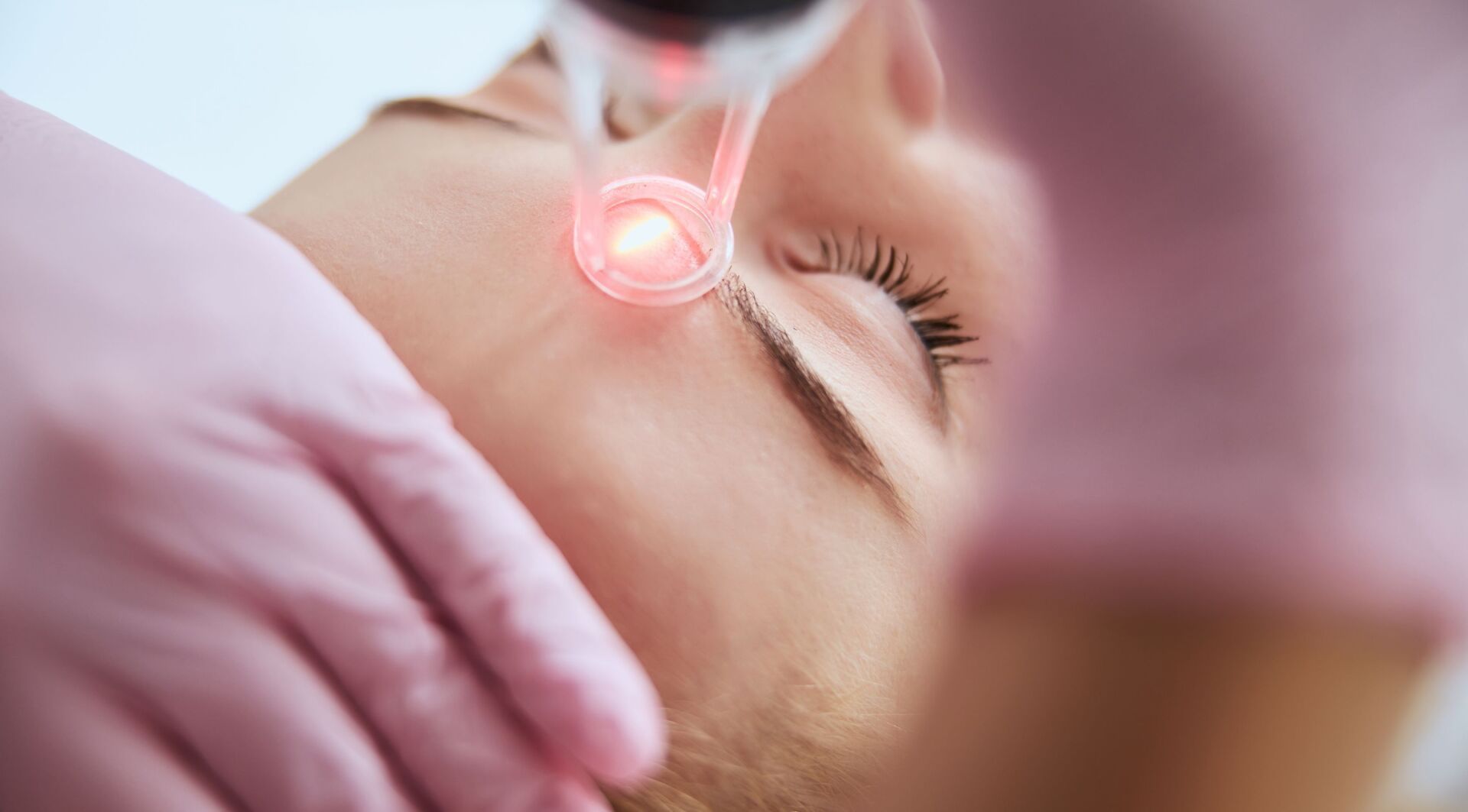 Person receiving a facial treatment with a red light device near their forehead. Pink gloves visible.