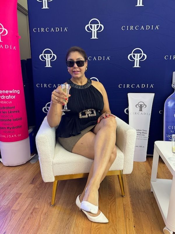Woman in sunglasses, holding champagne, seated in a white chair, Circadia backdrop.