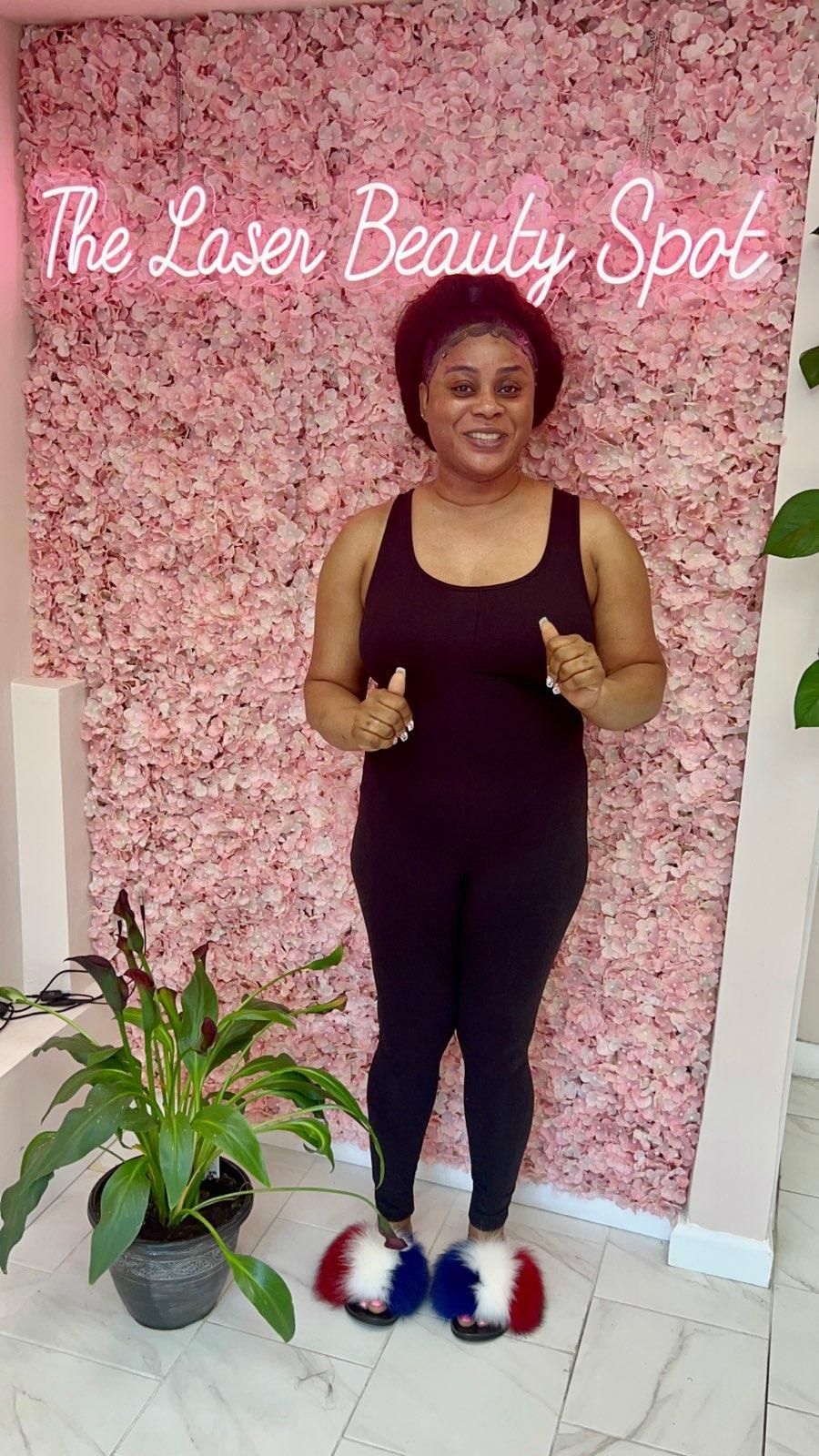 Woman in black outfit and fluffy slippers in front of a pink flower wall and neon sign.