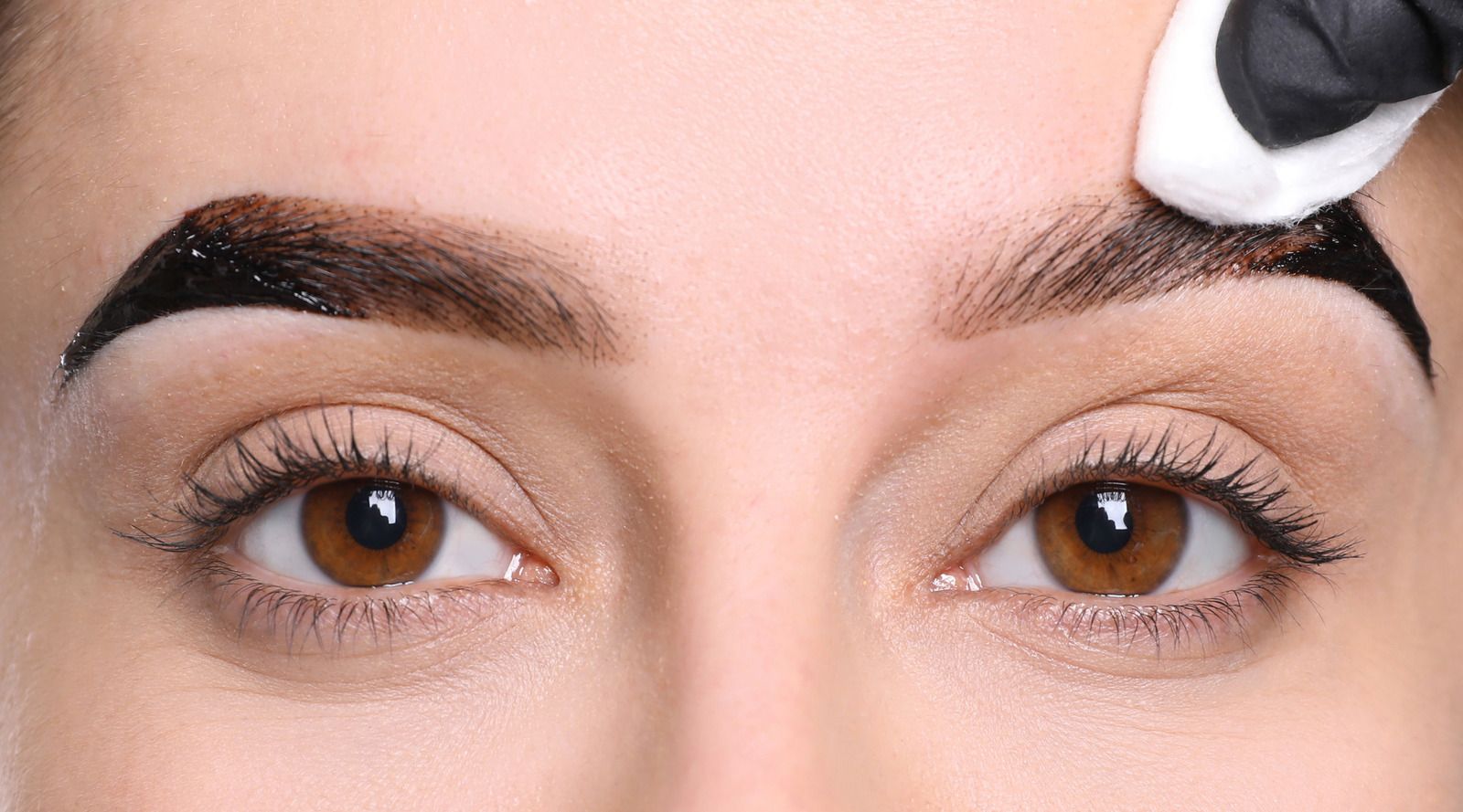 Eyebrow tint application with a gloved hand wiping excess dye. Brown eyes, close-up.