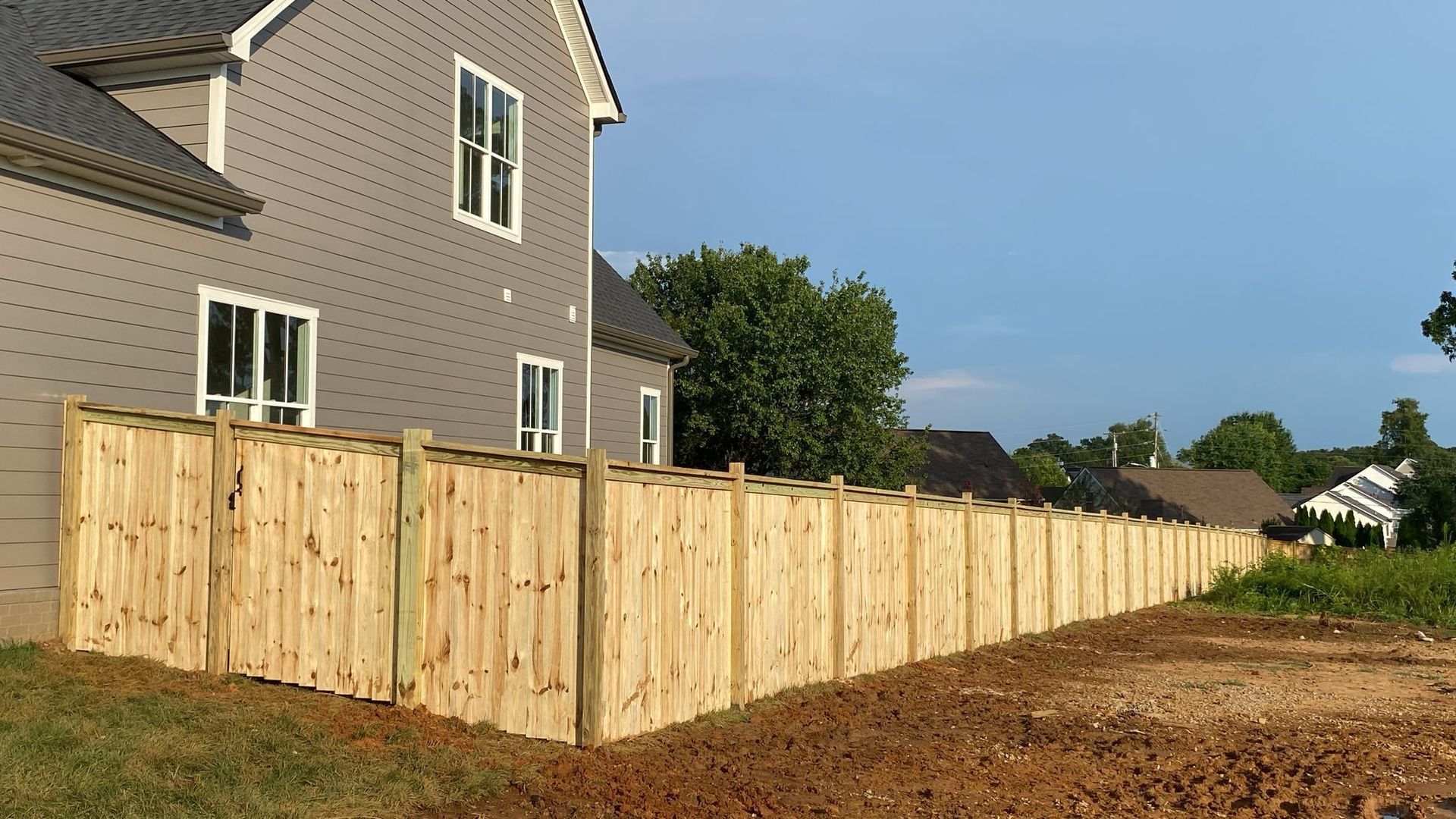 Wooden fence bordering a two-story gray house under a clear sky. Bare earth in front.