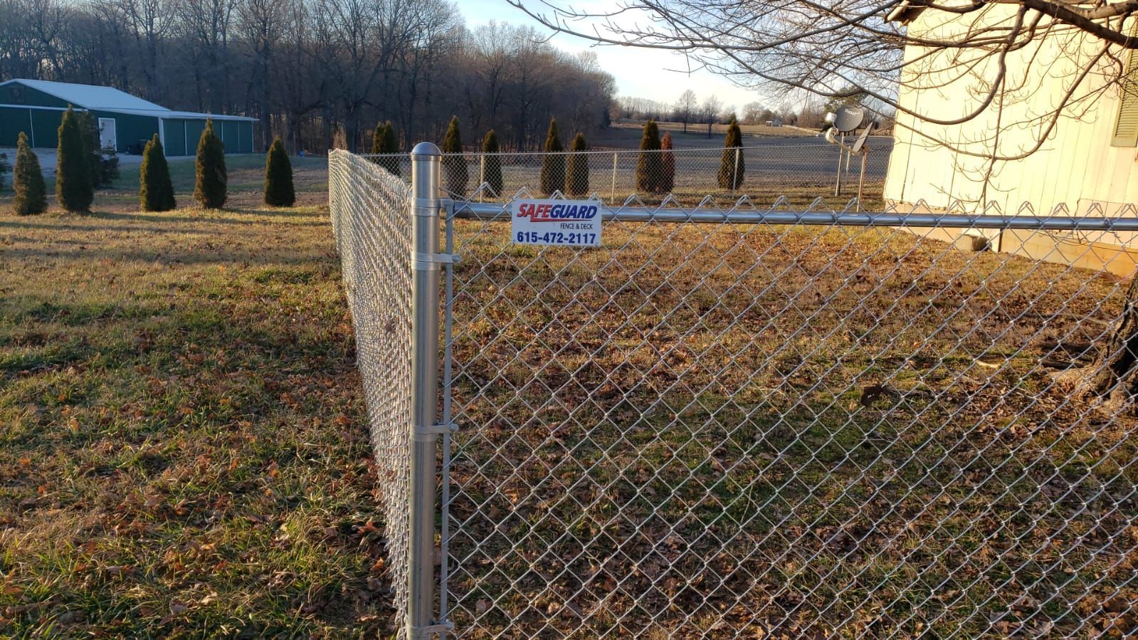 Chain-link fence in a grassy field with a sign. Trees and a building are in the background.