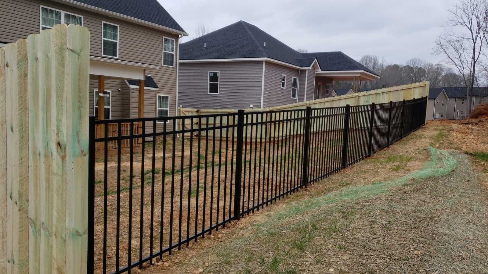 Black metal fence and wooden structure in front of new houses under construction. Gravel path in the foreground.