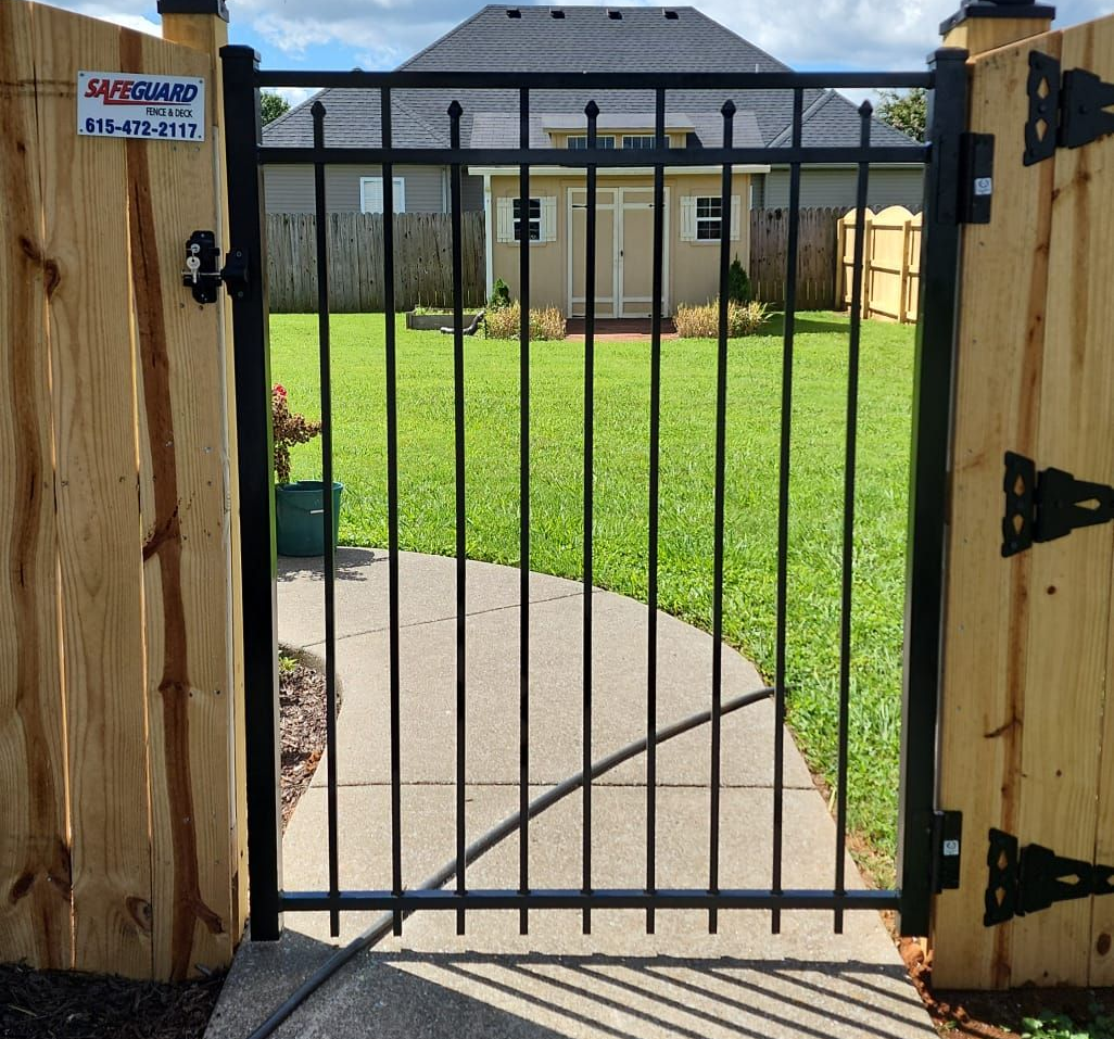 Black metal gate in a wooden fence, leading to a green lawn and house.