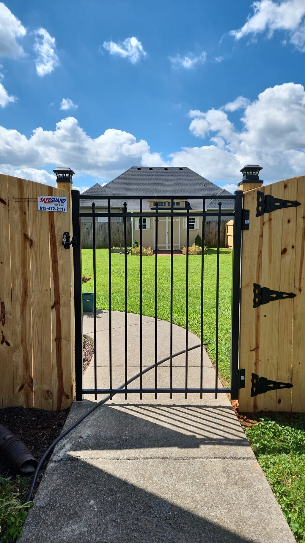 Open gate in a wooden fence leads to a backyard with grass and a house under a blue sky with clouds.