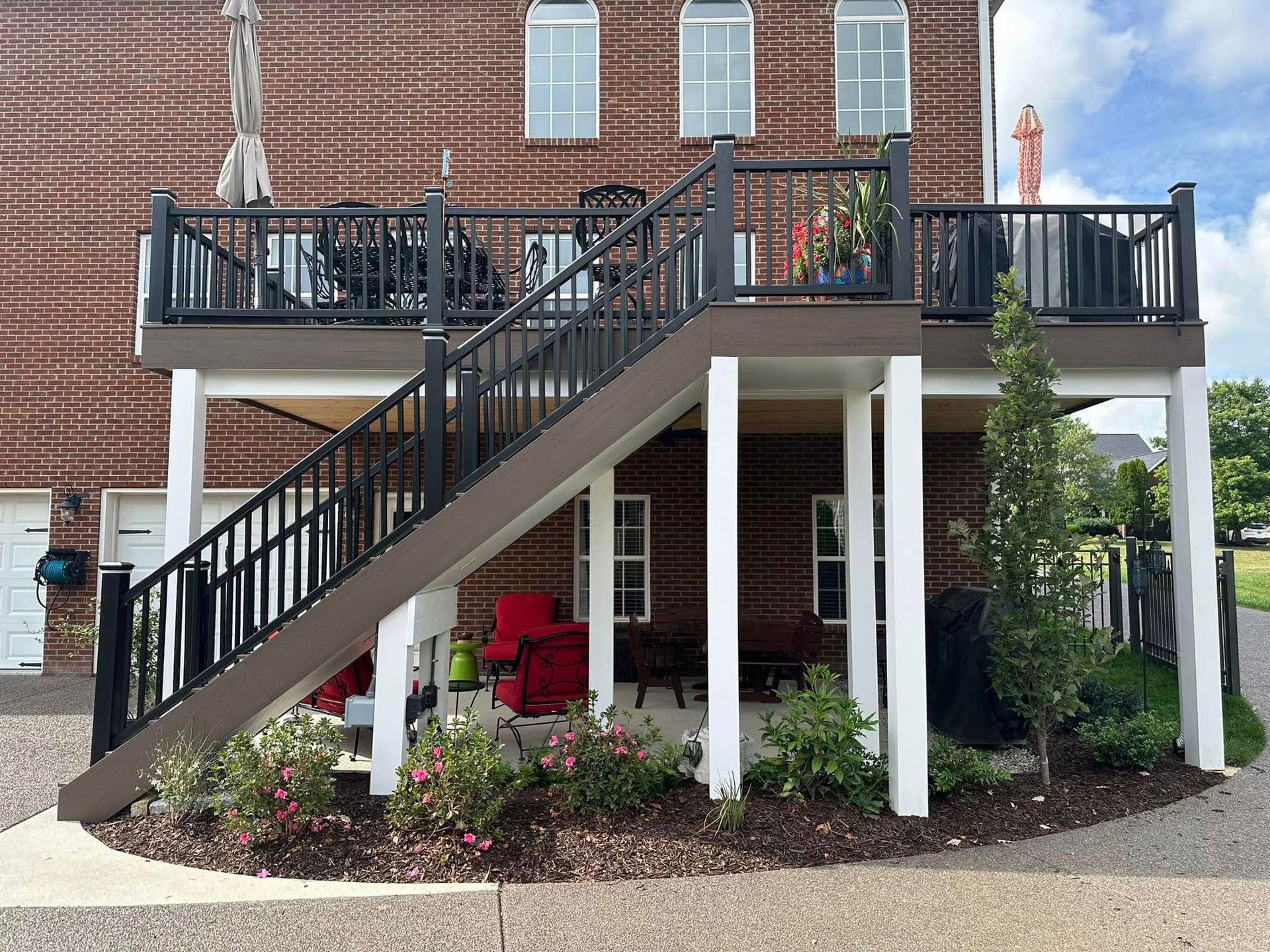 Two-story deck with stairs. Brick house, black railing, brown decking, white support posts, and small garden below.