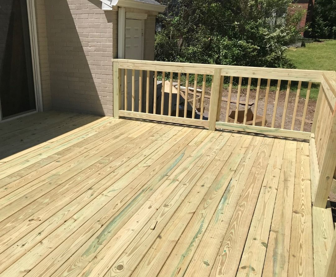 Newly built wooden deck with railing, connected to a light-colored brick house and lush greenery.