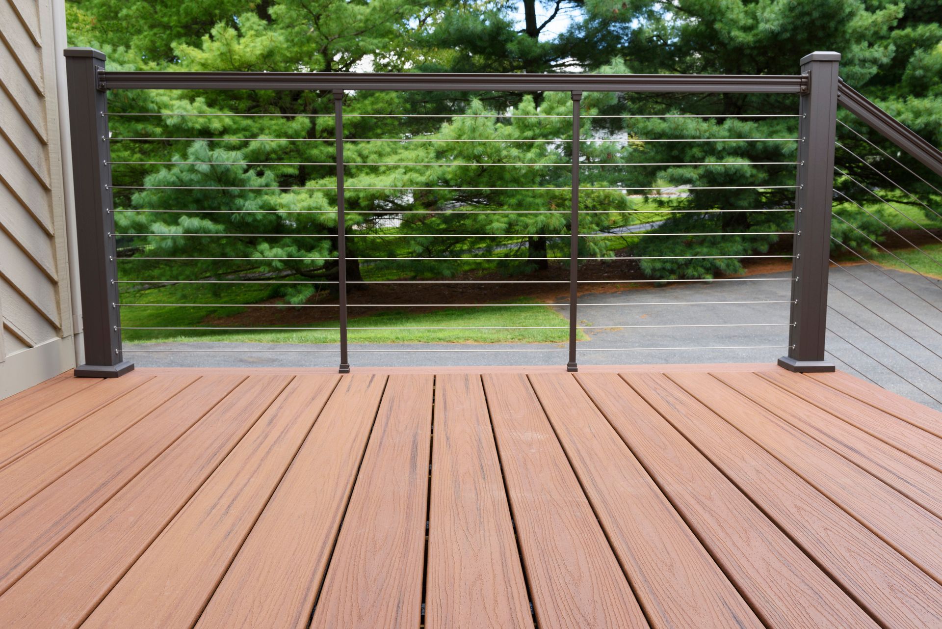 Brown deck with metal railing and cable infill, overlooking a driveway and trees.
