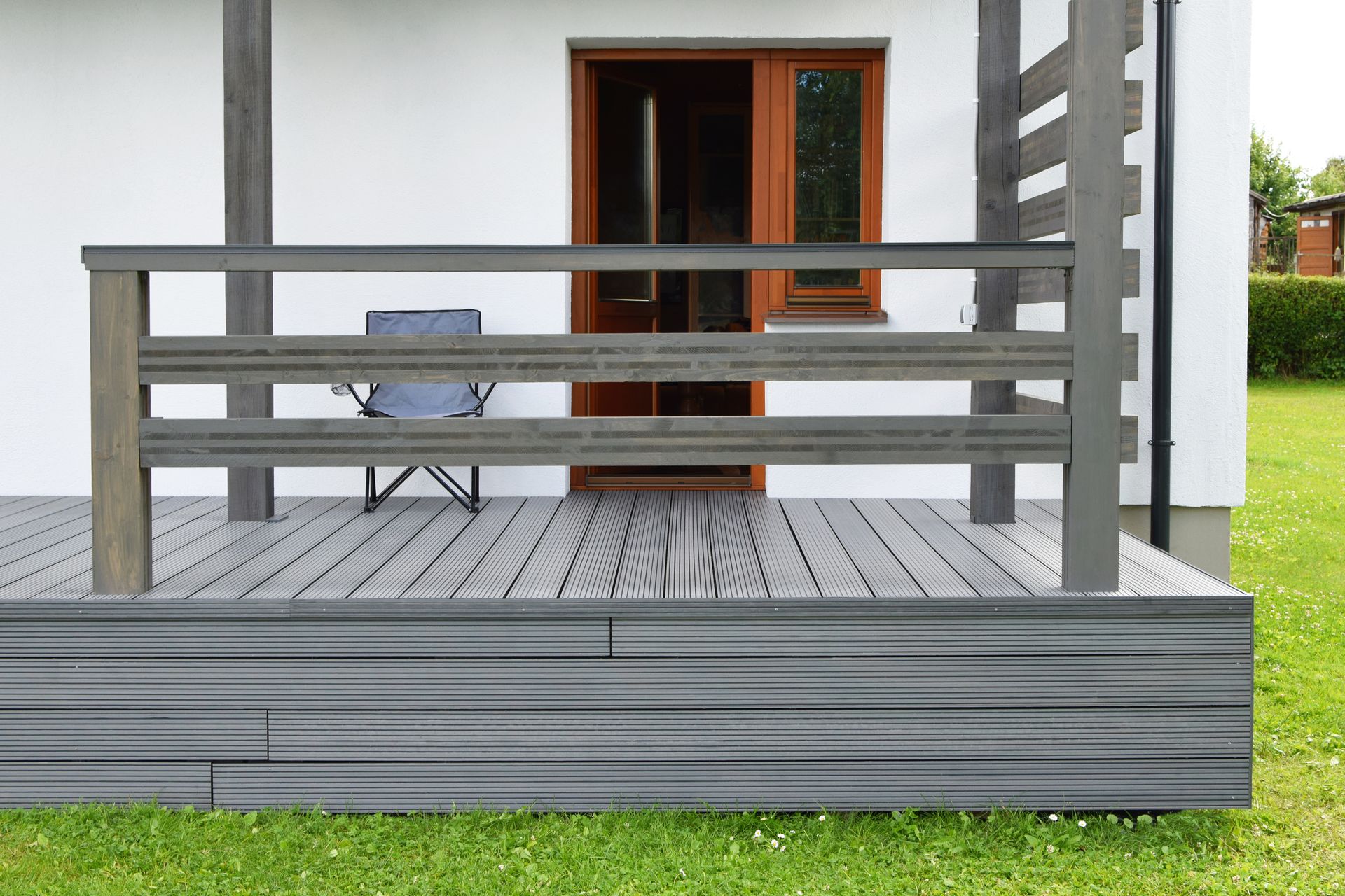 Gray composite deck with matching railing, chair near brown door of a white building, set in green grass.