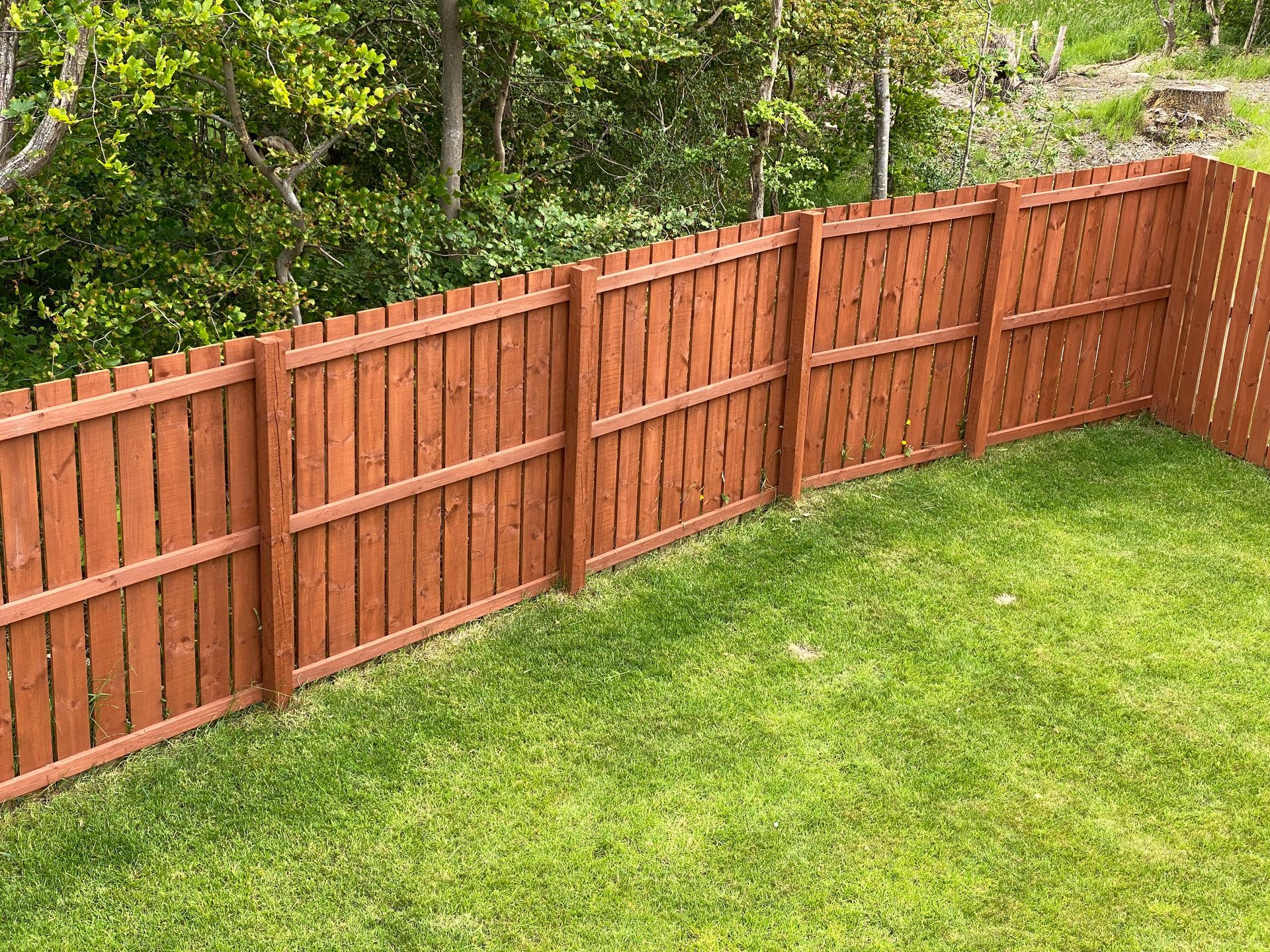 Wooden brown fence enclosing a green grass yard, with trees in the background.