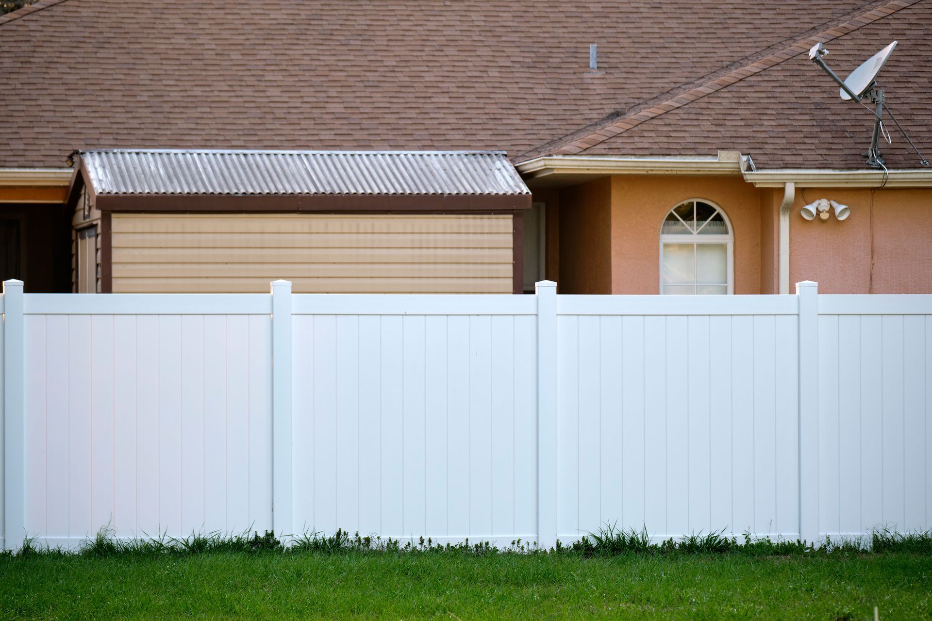 White fence in front of a tan shed and house with a satellite dish on a brown roof.