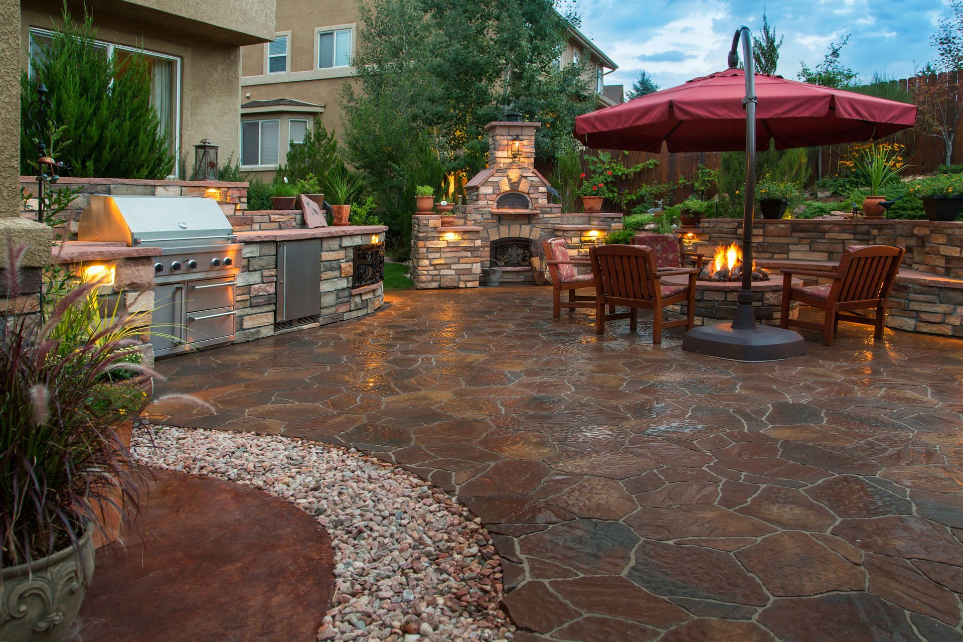 Outdoor patio with built-in grill, fireplace, seating, and umbrella; stone flooring and a gravel pathway.