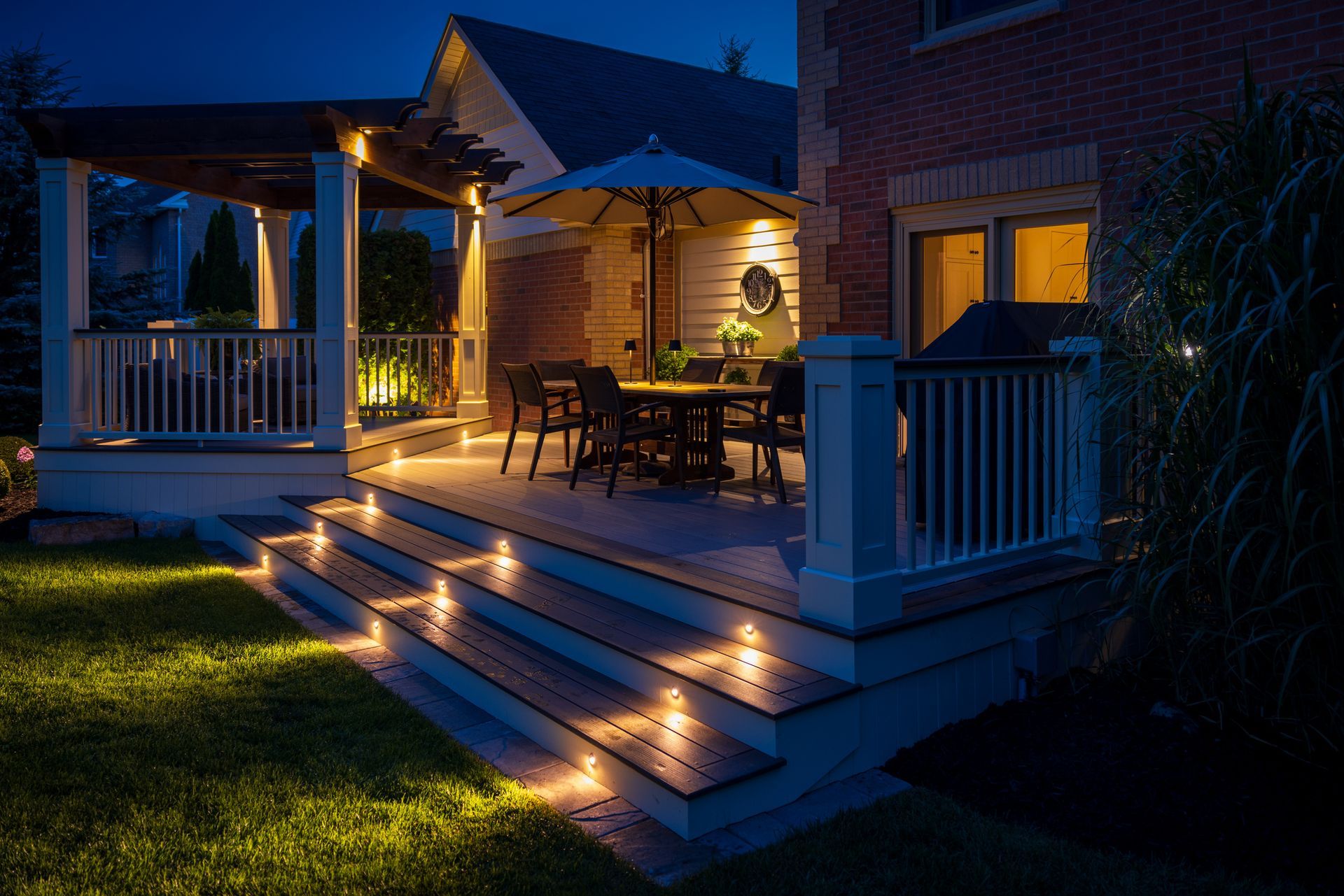 Lit outdoor deck at night with steps and pergola. Dining set under umbrella.