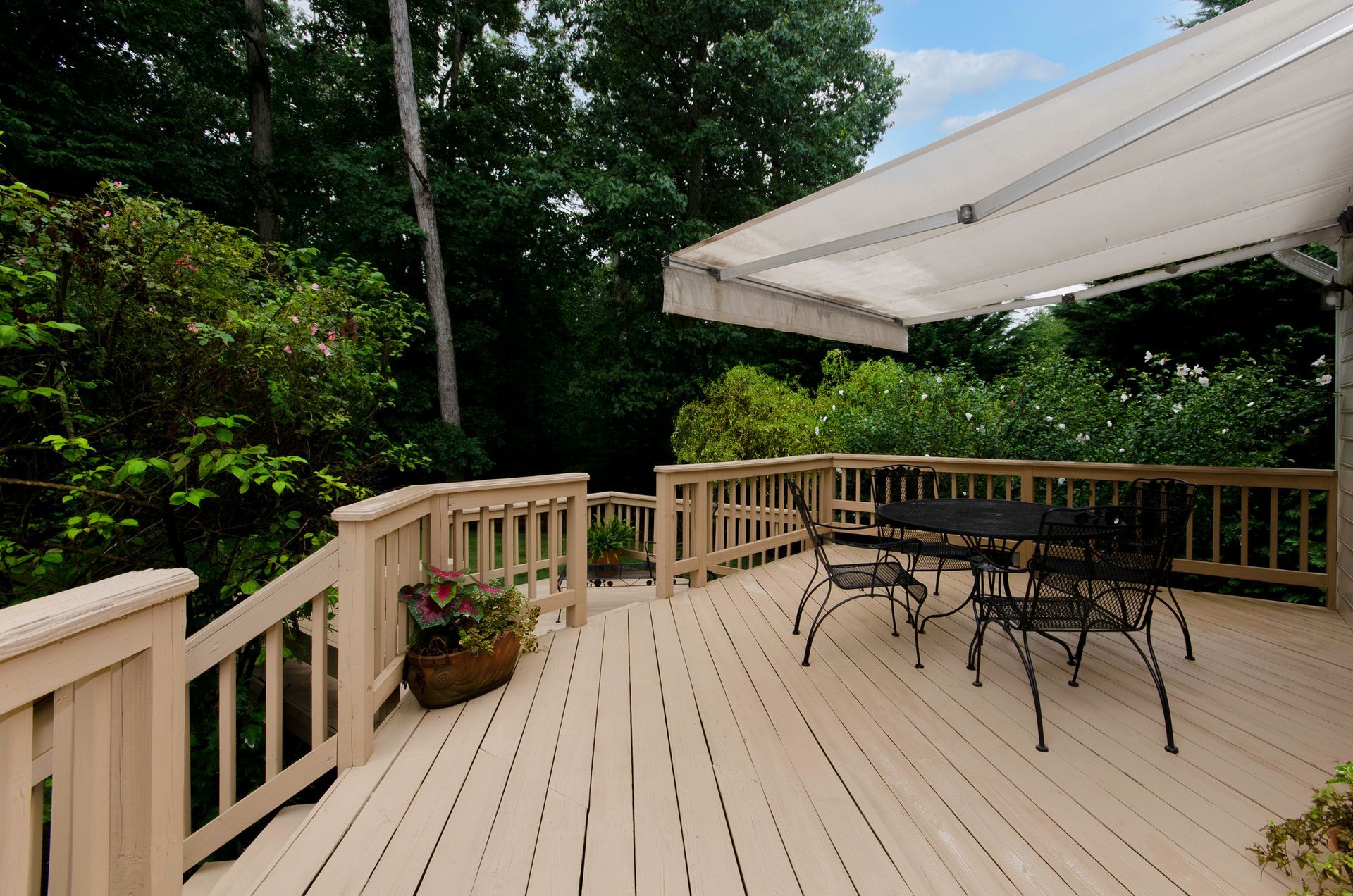 Wooden deck with black table and chairs, overlooking lush greenery and a white awning.