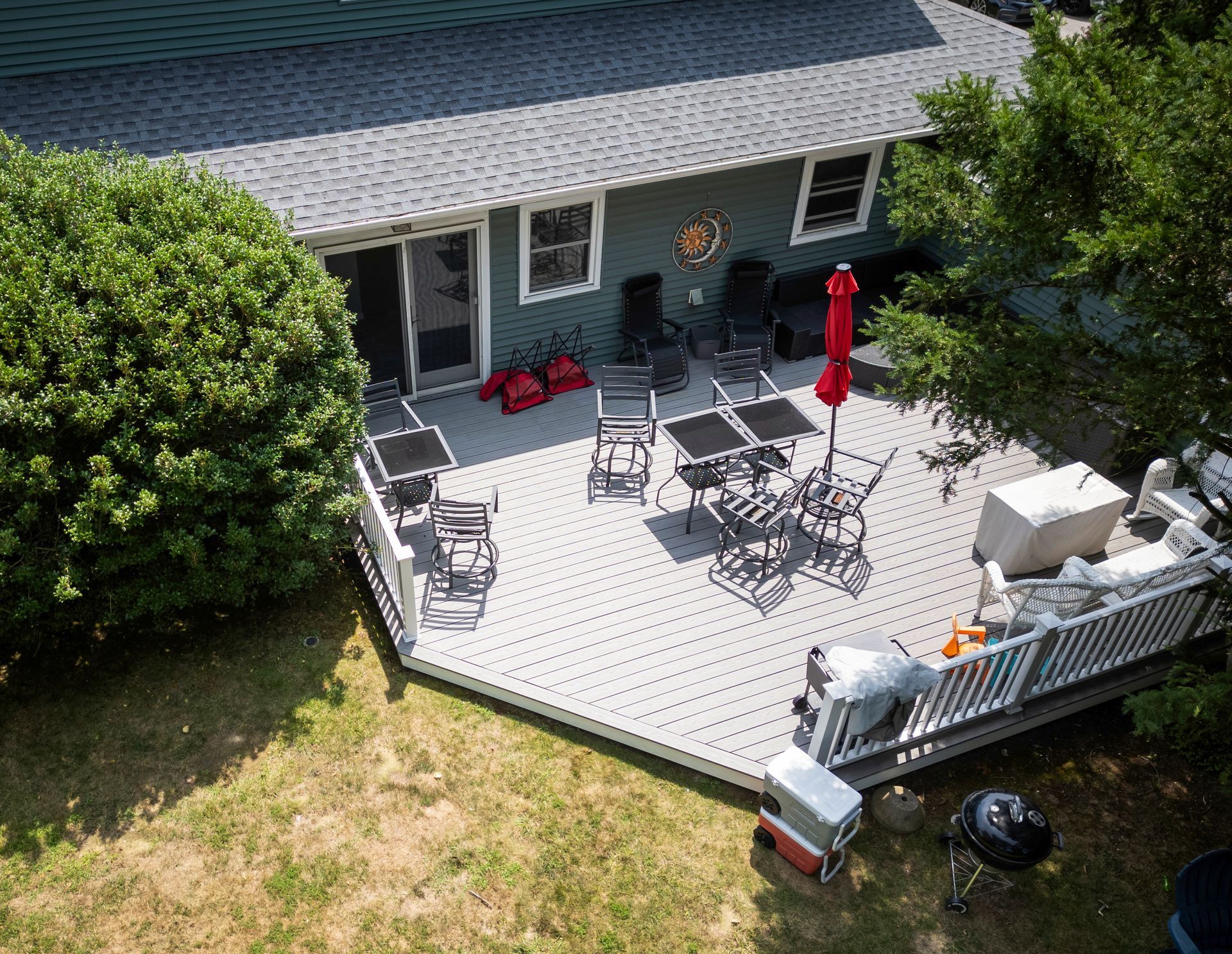 Overhead view of a gray deck with tables and chairs outside a green house, with lawn and shrubs surrounding.