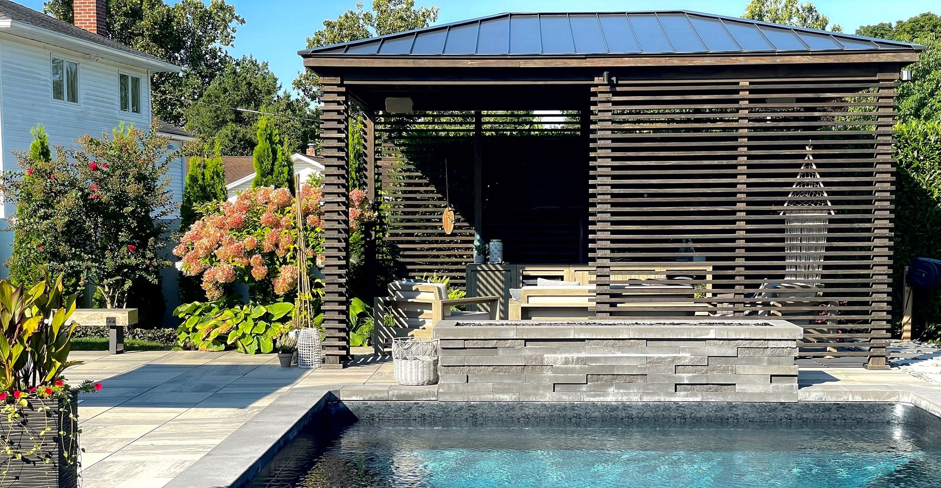 Poolside gazebo with dark wooden slats, pool, stone patio, and green trees.