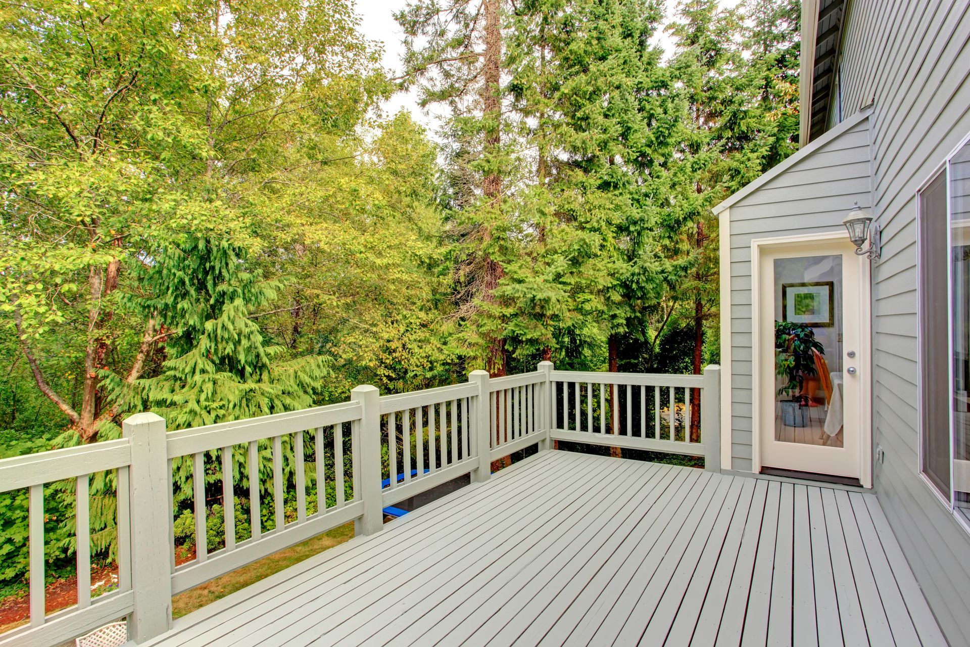 Wooden deck with white railing next to trees and a glass door.
