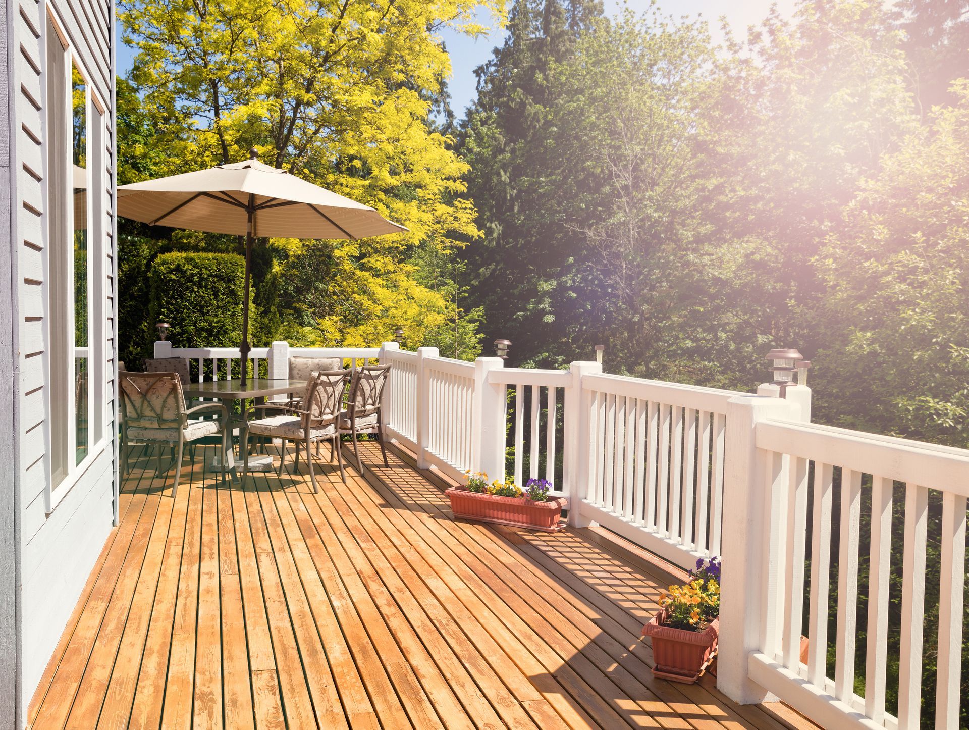 Wooden deck with white railing, table, chairs, and umbrella, with sunlight filtering through surrounding trees.