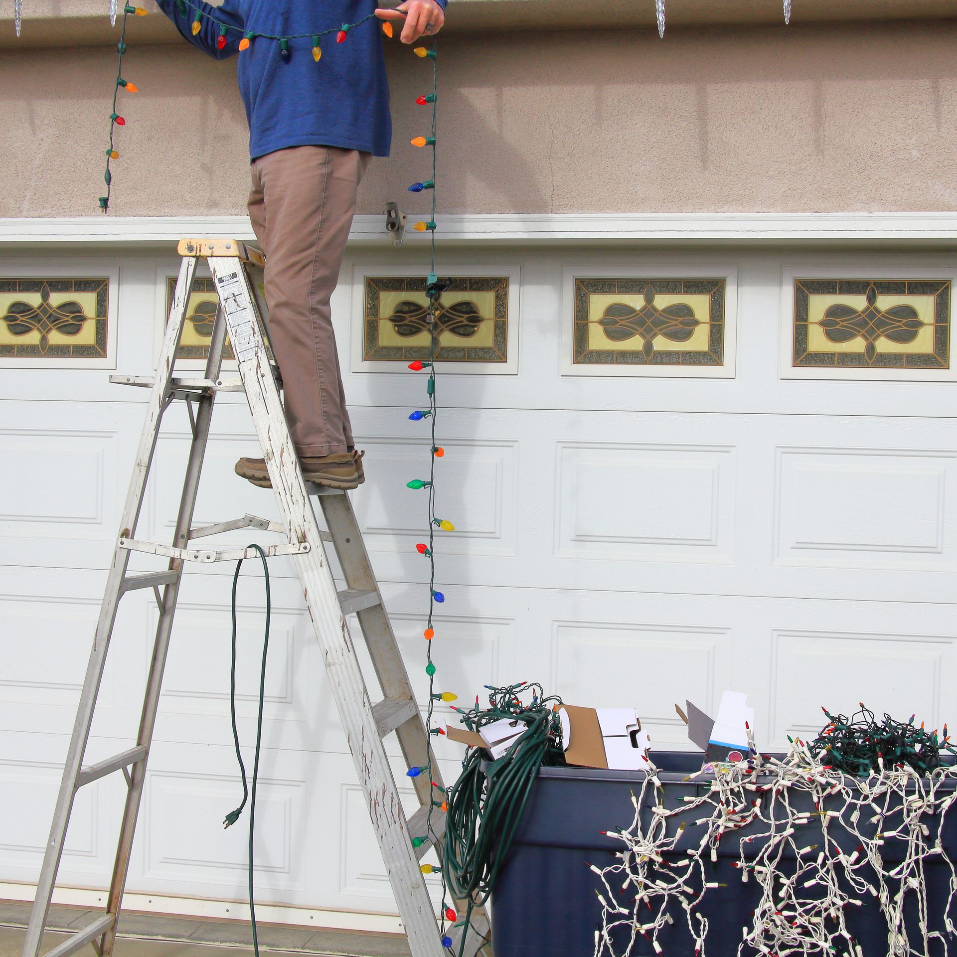 Person on ladder hanging colorful Christmas lights on garage.
