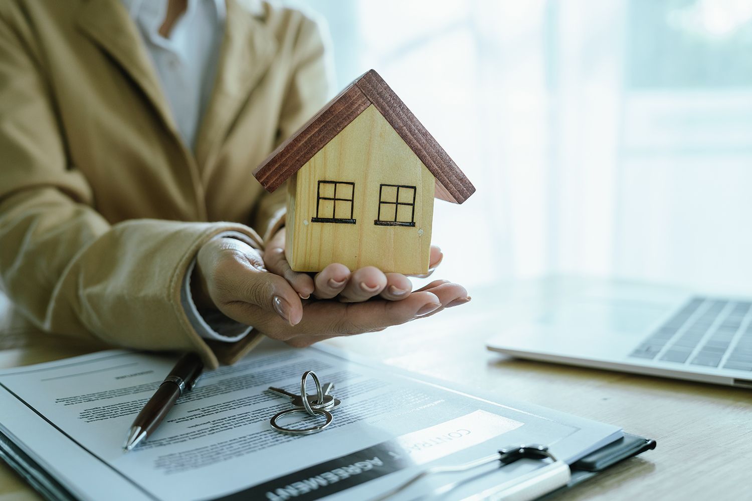 Person in a blazer holds a small wooden house over a contract and keys. Person in a blazer holds a small wooden house over a contract and keys.