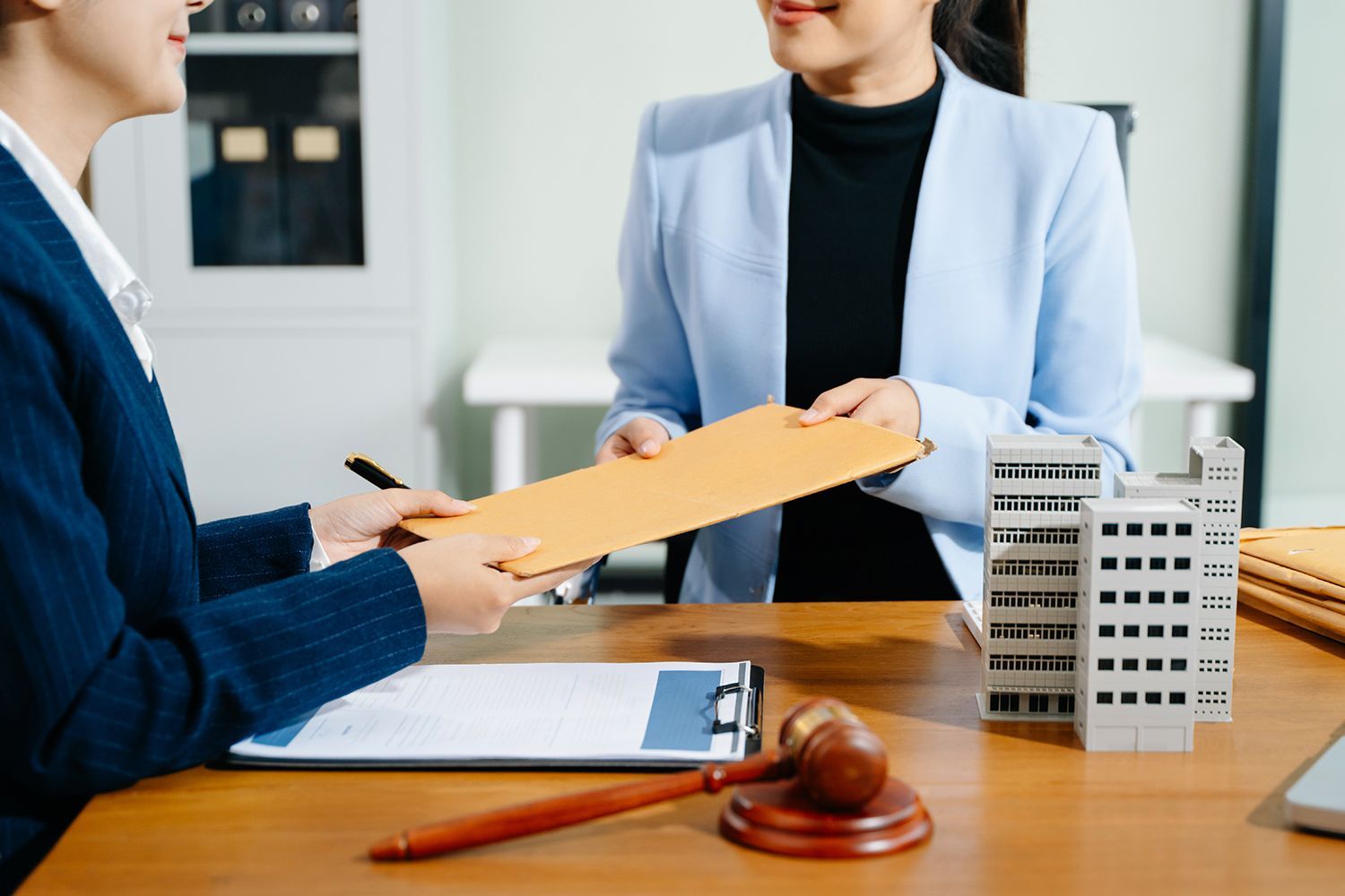 Two people exchanging documents at a desk, gavel and building models present.