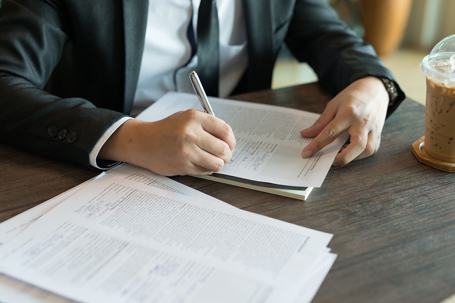 Person in a suit writing on papers at a table with a drink nearby. Person in a suit writing on papers at a table with a drink nearby.